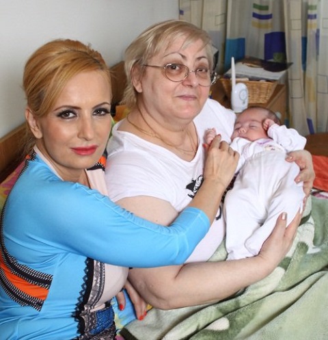 Daniela Gyorfi cu parul strans in coada, imbracata in rochie turcoaz mulata, stand fericita langa mamei si tinand manuta fiicei sale, Maria, in timp ce micuta este tinuta in brate de bunica ei, care poarta ochelari, tricou alb si sta in pat, rezemata de perne