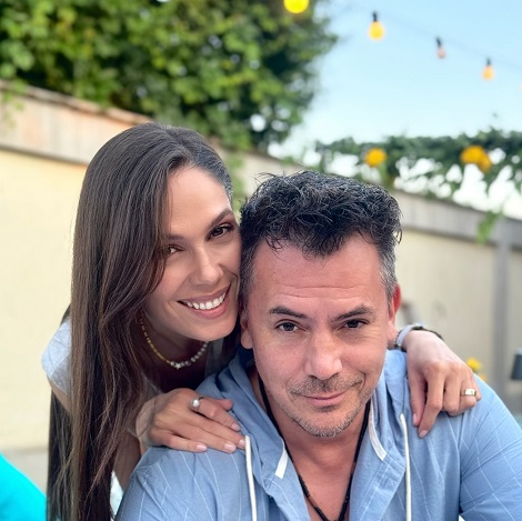 Irina Fodor in bluza fara maneci, cu colier scurt la gat,fericita in timp ce il cuprinde pe Razvan Fodor de umeri, acesta fiind imbracat cu un hanorac bleu cu dungi fine albe, amandoi stand in aer liber, in apropierea unui zid