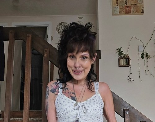 Theresa McIntosh vesela, avand un tatuaj cu floare in zona pieptului,imbracata in rochie alba cu imprimeu marunt gri, decoltata si cu bretele subtiri, stand rezemata de scara de lemn din locuinta ei, in spatele ei observandu-se pereti albi cu un tablou maro