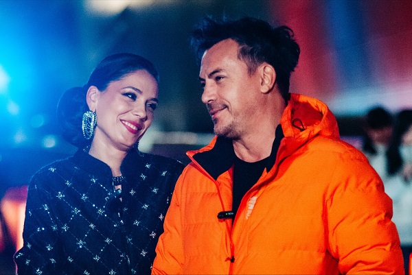 Irina Fodor imbracata cu sacou negru de catifea cu imprimeu argintiu, cu cercei argintii mari, fericita in timp ce il priveste pe sotul ei, Razvan Fodor, imbracat in geaca portocalie, pe platourile unei emisiuni TV