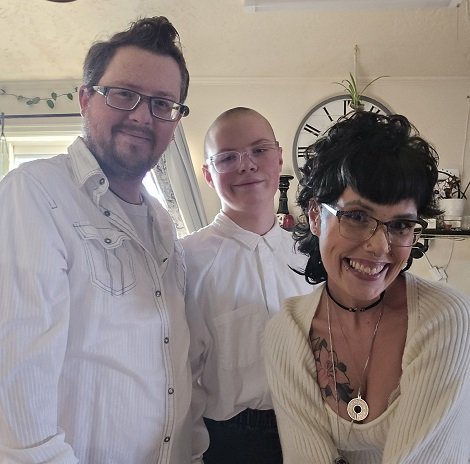 Theresa McIntosh fericita, imbracata in alb, cu ochelari, stand aplecata langa fiul si langa sotul ei, amandoi purtand ochelari si fiind imbracati in camasi albe cu maneca lunga