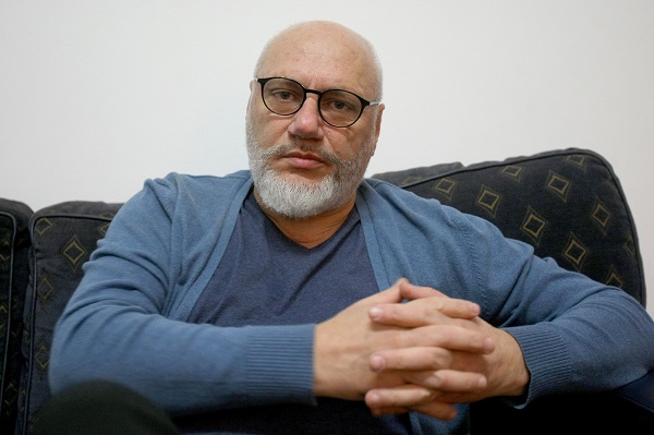 Cristian Andrei imbracat cu bluza si pulover albastre, purtand ochelari si stand ganditor pe o canapea bleumarin cu modele in forma de romb, in timp ce isi tine palmele impreunate 