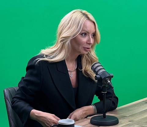 Daciana Sarbu imbracata in sacou negru, pe fundal verde, in timp ce sta pe un scaun negru de piele, la o masa, si vorbeste la un microfon pus pe masa la o emisiune si are langa ea o cana neagra cu alb