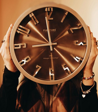 femeie imbracata cu bluza neagra cu maneci lungi si cu vesta maro matlasata, avand doua bratari la una dintre maini si tinand in maini un ceas mare cu cadran maroniu ascuzandu-si fata