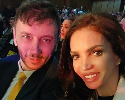 Cristina Spatar imbracata cu sacou alb si bluza neagra fericita alaturi de sotul ei, Vicentiu Mocanu, imbracat in sacou inchis la culoare, camasa alba si purtand cravata galbena, in timp ce se afla amandoi intr-o sala plina la un spectacol