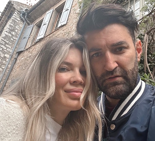 Gina Pistol zambitoare, imbracata in alb, in timp ce isi sprijina capul de Smiley, imbracat cu o jacheta bleumarin cu dunga alba, in timp ce in spatele lor se afla o vila cu peretii de piatra pe o straduta dintr-o localitate din Franța