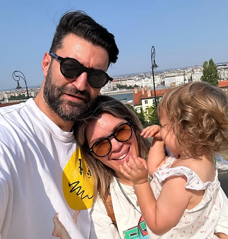 Smiley purtand ochelari de soare, imbracat cu tricou alb cu inscriptie galbena,fericit alaturi de Gina Pistol, imbracata tot in tricou alb, cu scris colorat, aceasta tinand-o in brate pe fiica lor, imbracata cu rochita cu bretele, cu spatele la camera, in timp ce se afla in vacanta vizitand un oras din strainatate