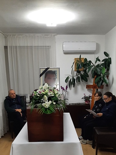 imagine de la funeraliile lui George Romila, romanul de 46 de ani gasit mort in Spania, sicriu maro asezat pe masa, cu flori albe deasupra, un barbat in varsta in negru stand pe un scaun si doua femei in varsta, in haine inchise la culoare, stand langa sicriu, pe scaune, langa ele fiind plante exotice si crucea de lemn cu numele barbatului decedat