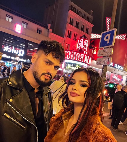 influencerita Andreea Ciuca zambitoare, imbracata cu bluza maro si cu jacheta de blana asortata, stand langa fostul ei iubit, imbracat cu jacheta neagra de piele, pe o strada aglomerata intr-o zona cu reclame luminoase din Paris