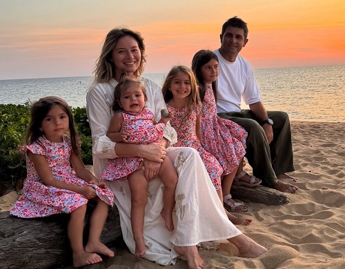 Laura Cosoi fericita, imbracata in rochie alba lunga, tinandu-si in poala fetita cea mica, in timp ce sta, alaturi de celelalte trei fetite, imbracata toate in rochite cu flori roz, si alaturi de sotul ei, Cosmin Curticapean, imbracat in tricou alb si pantaloni maro, pe o plaja, la apusul soarelui, in vacanta din Thailanda