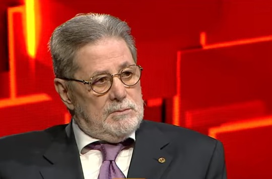 Cornel Dinu ganditor, purtand ochelari, imbracat in sacou negru, camasa alba si cravata violet, stand pe scaun in timpul unei emisiuni si avand in spatele sau fundal roșu