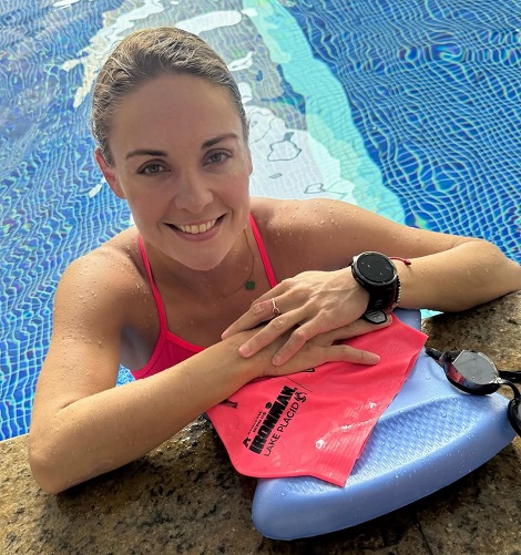 Mara Flavia Araujo vesela, cu parul strans si ud, in costum de baie rosu in piscina, stand cu coatele sprijinite de marginea piscinei, avand langa ea o pluta cu simbolul competitiei IRONMAN