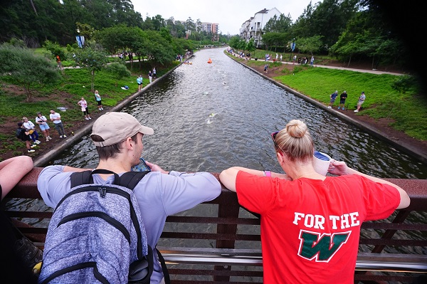 femeie imbracata in tricou rosu si barbat imbracat cu tricou gri, purtand sapca bej si avand in spate un rucsac albastriu in timp ce amandoi stau cu spatele pe un pod si se uita la o apa pe care se desfasoara competitia de inot Texas Ironman, pe marginile apei fiind si alte persoane care urmaresc competitia 