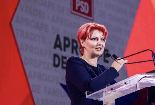 Olguta Vasilescu imbracata in rochie bleumarin, stand la pupitru si tinand cu mana de un microfon in timpul unei sedinte a PSD