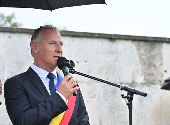 Dinu Iancu-Salajanu imbracat in costum inchis la culoare, cu camasa alba si cravata bleumarin, avand pe sacou o banderola cu tricolorul, tinand in mana un microfon si luand cuvantul la o reuniune in aer liber, intr-o zi ploioasa, fiind protejat de o umbrela neagra