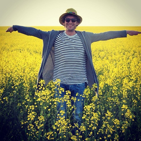 Mircea Solcanu fericit, cu palarie pe cap, cu ochelari negri de soare, imbracat cu tricou alb cu dungi negre, cu hanorac gri si blugi, in timp ce isi intinde bratele entuziasmat intr-un lan de rapită pe un c&acirc;mp