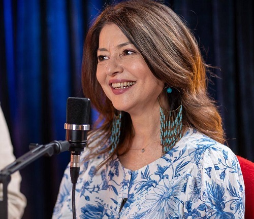 Oana S&icirc;rbu vesela, imbracata in rochie alba cu flori albastre, purtand cercei lungi turcoaz, in timp ce sta pe un scaun rosu in fata unui microfon cand a fost invitata la un podcast 