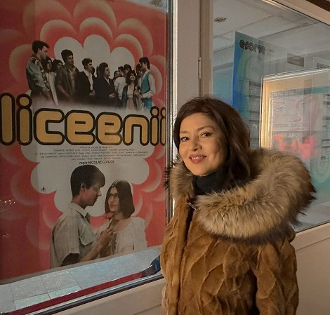 Oana S&icirc;rbu zambitoare, imbracata intr-o haina de blana maronie, cu guler bej, in timp ce sta in fata unui panou de sticla pe care este lipit un afis cu filmul Liceenii
