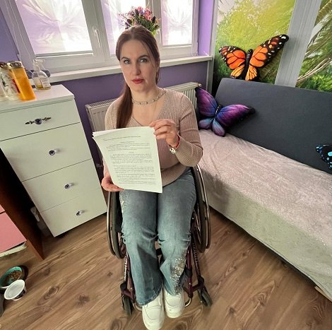 Crina Steliana Tugui, membru USR, imbracata in bluza roz pal si blugi, tinand in mana un document, in timp ce sta in scaunul ei cu rotile langa o canapea gri decorata cu perne fluture, intr-o camera cu peretii lila