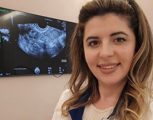 medicul Cristina Buzgar, zambitoare, imbracata in bluza crem si in halat crem cu guler albastru inchis, in timp ce sta cu spatele la un monitor pe care se vede imaginea provenita de la o ecografie detala
