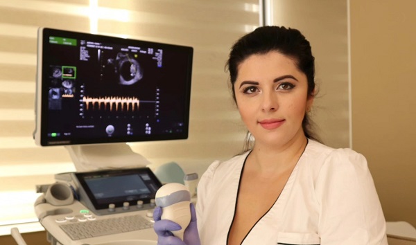 medicul ginecolog Cristina Buzgar, imbracata in halat alb, stand pe scaun si tinand in mana cu manusi chirurgicale mov un dispozitiv pentru ecografia fetala, in timp ce sta pe scaun, cu spatele la ecograful dintr-un cabinet medical