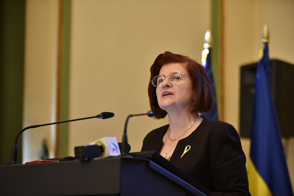 dr. Catalina Poiana, presedintele Colegiului Medicilor din Romania, imbracata in costum negru, purtand ochelari si o brosa aurie, in timp ce sta la un pupitru cu microfoane pentru o interventie la un eveniment