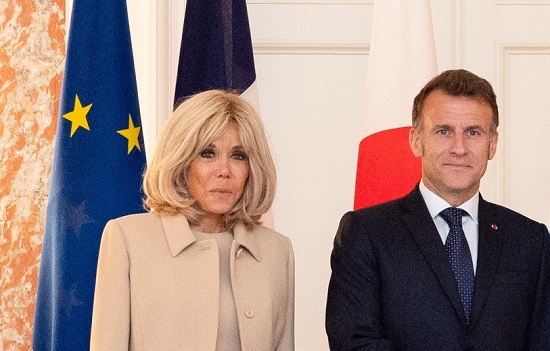 Brigitte Macron imbracata in costum bej, alaturi de sotul ei, Emmanuel Macron, imbracat in costum bleumarin, camasa alba si purtand cravata bleumarin cu buline albe, amandoi aflandu-se intr-o vizita oficiala 