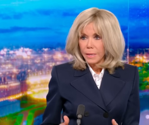 Brigitte Macron imbracata cu sacou bleumarin si camasa alba, stand pe scaun, in fata microfonului, in timpul unei interventii la un post de televiziune