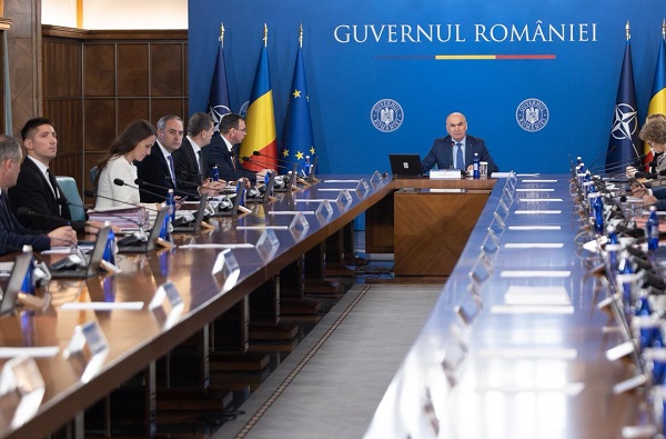 Ilie Bolojan, imbracat in costum bleumarin, cu camasa bleu si cu cravata bleumarin, conducand o ședintă de Guvern la care participa mai multi ministrii