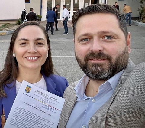 Anamaria Gavrila vesela, imbracata cu sacou albastru si bluza alba, tinand in mana documente eliberate de Biroul Electoral Central, in timp ce sta langa Razvan Chirita, imbracat cu sacou alb cu gri cu patratele si cu camasa bleu, care face selfie, in spatele lor aflandu-se mai multe persoane care pregătesc o secție de votare