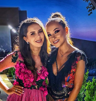 Georgiana Lobonț veselă, îmbracată cu rochie ciclam cu negru, tinandu-si una dintre maini in sold, in timp ce se afla alaturi de nasa ei, Vladuta Lupau, imbracata cu rochie neagra cu ornamente stralucitoare de diverse culori intr-un spatiu in aer liber seara, la un eveniment