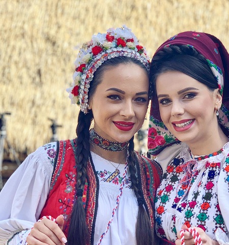 Vlăduța Lupău și Georgiana Lobonț vesele, îmbrăcate amândouă cu costume populare, stand impreuna într-un spatiu din aer liber si tinand fiecare in mana cate un snur de martisor