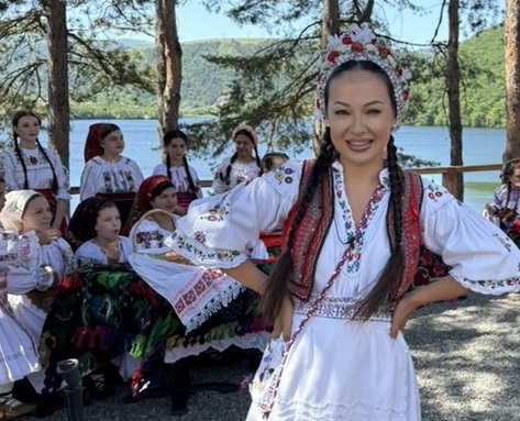 Vlăduța Lupău in costum popular, tinandu-si mainile in solduri, in timp ce este inconjurata de femei in costume populare la un eveniment in aer liber, intre copaci, pe malul unei ape