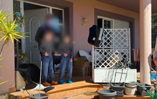 captură video, persoana adulta alaturi de doi baieti, imbracati la fel, cu bluze gri cu dungi negre si pantaloni albastre care stau pe treptele unei case vopsită roz pregatindu-se sa iasa in curtea plina de diverse obiecte, imagine din casa din Oviedo, Spania, &icirc;n care trei copii au fost ținuți &icirc;nchiși &icirc;n casă de părinții lor 4 ani