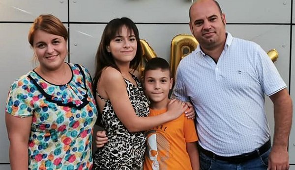 Alexandra Căpitănescu fericita, imbracata intr-o rochie cu bretele cu animal print imbratisandu-si fratele, Radu Capitanescu, imbracat cu tricou galben, amandoi avandu-i alaturi pe parintii lor, mama imbracata cu bluza pe fond alb cu buline colorate si tatal, Gabriel Căpitănescu, imbrăcat cu tricou cu imprimeu marunt si blugi