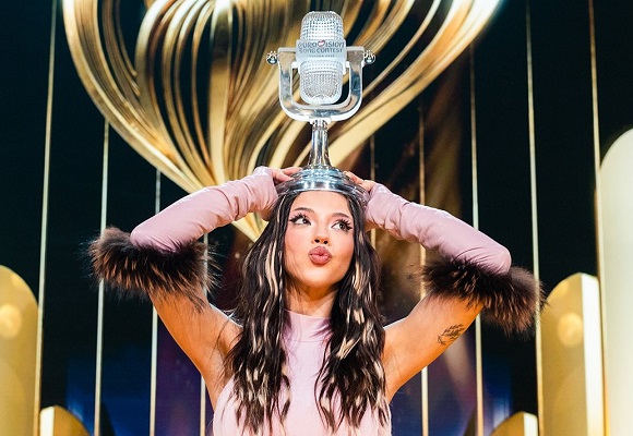 DARA, câștigătoare Eurovision 2026, imbracata in roz, cu ornamente de blana la maneci, tinand pe cap trofeul Eurovision, aflandu-se pe scena dupa marea finala, in spatele ei putandu-se vedea simbolul Eurovision, cu auriu