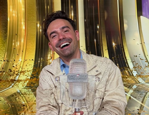 Compozitorul si producatorul muzical Cristian Tarcea, imbracat cu costum crem si camasa bleu, fericit, in timp ce se afla pe scena Eurovision, cu confetti aurii, si tine in mana trofeul Eurovision