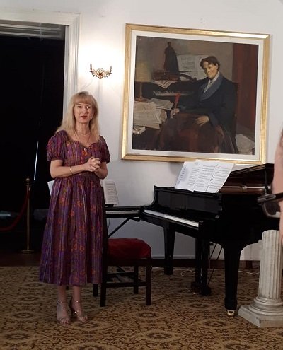pianista Alina Pavalache imbracata cu rochie visinie cu flori, stand langa un pian intr-o sala cu un tablou care intruchipeaza un compozitor in timp ce tine un discurs