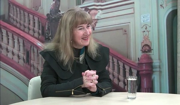 pianista Alina Pavalache vesela, imbracata cu helanca si cu jacheta neagra, tinandu-si palmele stranse in timp ce statea pe un scaun, la o masa alba pe care se afla un pahar cu apa, in timp ce era invitata la o institutie 