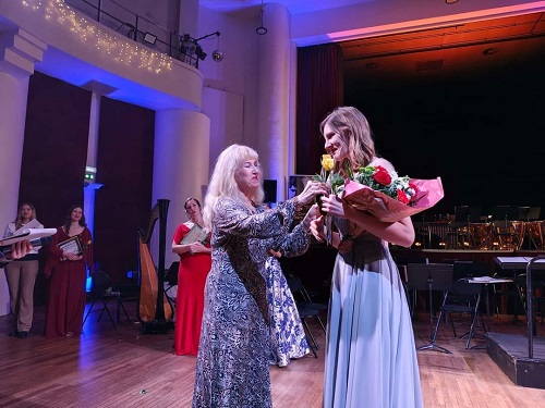 pianista Alina Pavalache, imbracata cu rochie alba cu imprimeu negru, in timp ce se afla pe o scena si ii daruieste o floare unei tinere imbracate in rochie rosie, care a participat la un spectacol sau la un concurs, in timp ce in preajma lor se afla si alte tinere premiante