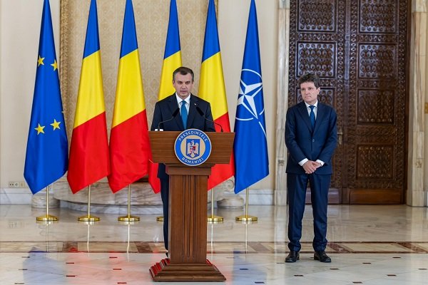 Eugen Tomac, premierul desemnat, imbracat cu costum bleumarin, cu camasa alba si cu cravata albastra, stand la pupitrul Presedintelui Romaniei, la Administratia Prezidentială, avand in spatele sau diverse steaguri, in timp ce presedintele Nicusor Dan, imbracat tot cu costum bleumarin sta deoparte, cu spatele la o usa de lemn masiv, după ce l-a desemnat premier