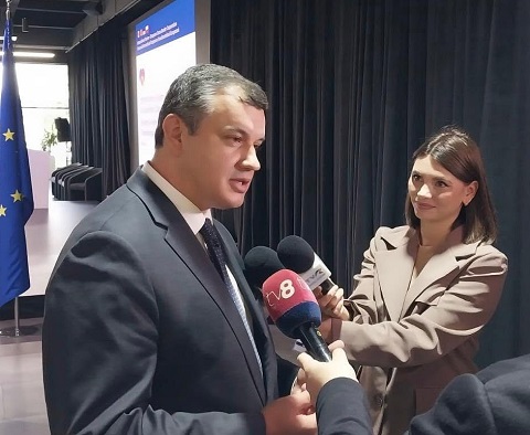 Premierul desemnat, Eugen Tomac, imbracat cu costum inchis la culoare, cu camasa alba si cravata, in timp ce acorda un interviu pentru doi reporteri, aflandu-se intr-o sala inainte sau dupa o intalnire oficiala, in spatele lui putandu-se observa steagul Uniunii Europene