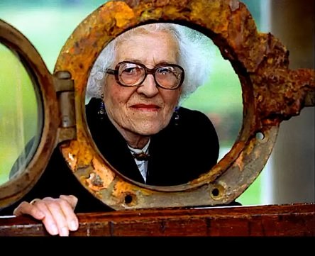 Millvina Dean, cea mai tanara pasagera de pe Titanic, purtand ochelari, fiind imbracata in sacou negru, in timp ce se afla la o expozitie sau la un muzeu legat de tragedia despre Titanic si sta cu capul incadrat intr-o fereastra recuperata de pe vas