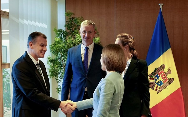 premierul desemnat Eugen Tomac, imbracat cu costum negru, cu camasa alba si cravata, zambitor in timp ce strange mana Maiei Sandu, președintele Republicii Moldova, imbracata cu costum vernil, in timpul unei &icirc;nt&acirc;lniri din Republica Moldova, langa cei doi aflandu-se si alti doi oficiali, iar &icirc;n spatele acestora din urmă steagul Republicii Moldova