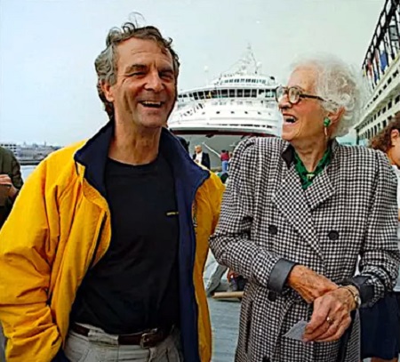 Millvina Dean, cea mai tanara pasagera de pe Titanic, imbracata cu o haina cu patratele alb-negru, cu maneci lungi, tinand in mana un bilet, in timp ce zambeste si se uita spre un barbat imbracat cu bluza neagra si jaceheta galbena, in timp ce amandoi se pregatesc sa urce la bordul unei nave de croaziera dintr-un port