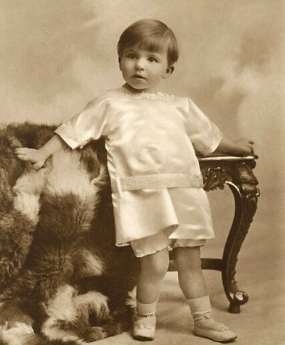 fotografie disponibila la Muzeul National Cotroceni, Principele Mircea, fiul Reginei Maria si al Regelui Ferdinand, imbracat cu un costumas alb, cu pantaloni scurti, in timp ce statea rezemat de un scaun vintage acoperit cu o blana