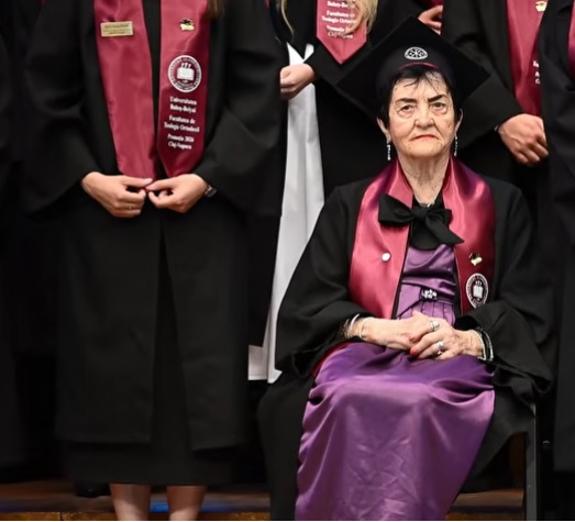 doamna Amalia-Susana Tușa, cea mai varstnica studenta din Romania, care a absolvitat facultatea la 90 de ani, imbracata cu rochie mov si cu roba, purtand toca pe cap in timp ce sta pe un scaun langa alti absolventi in timpul ceremoniei de absolvire a Facultatii de Teologie Ortodoxa din Cluj