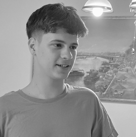 fotografie alb-negru, Dragos Florin Țucuina, adolescentul din judetul Timis care a murit in urma unui accident cu trotineta electrica,zambitor, imbracat cu tricou, avand in spatele sau un tablou