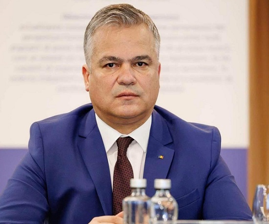 premierul desemnat Adrian Veștea, imbracat cu sacou albastru si camasa alba, avand gravata visinie, stand pe scaun la pupitru in timpul unei sedinte, in timp ce in fata lui sunt sticle de apă, iar in spatele sau se vede un ecran mare