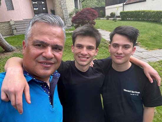 premierul desemnat Adrian Veștea imbracat cu tricou turcoaz, vesel, alaturi de baietii lui, imbracati amandoi cu tricouri negre, tinandu-si tatal dupa gat, in timp ce toti trei se afla in curtea casei